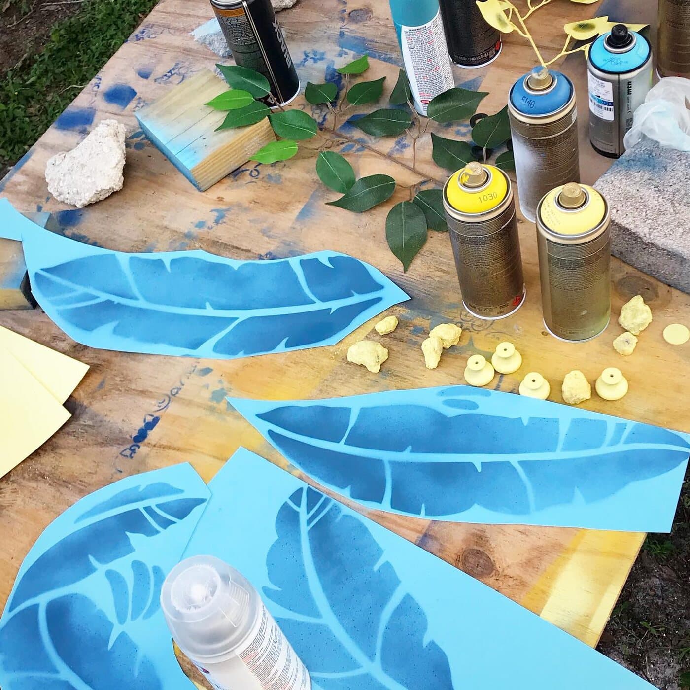 Stenciled blue leaves drying outdoors beside spray cans