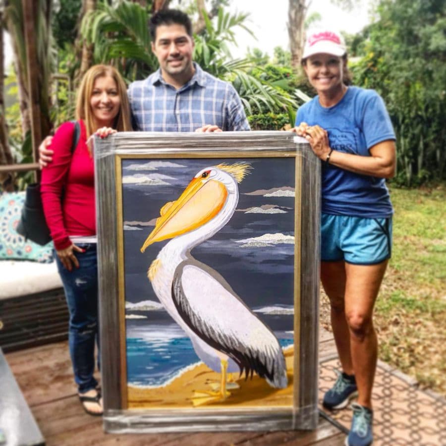 A couple and Jennifer with a framed white pelican piece under the trees