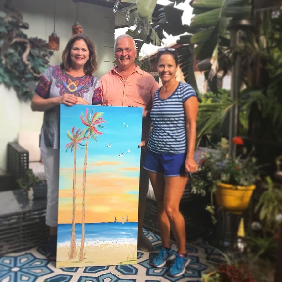 Three collectors with a tall palms-at-sunset canvas in a garden
